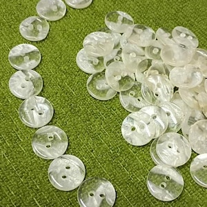 May include: A collection of clear, round buttons with two holes, arranged on a green textured surface. The buttons have a pearlescent, marbled appearance. Some buttons are arranged in a curved line, while others are in a pile.