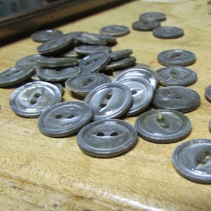 May include: A collection of gray, oval-shaped buttons with two holes each, scattered on a wooden surface. The buttons have a slightly iridescent sheen and varying shades of gray. Some buttons are in focus, while others are blurred.