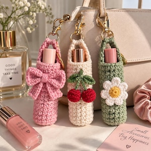 May include: Three crochet lipstick holders with gold-tone lobster clasps. The pink holder features a bow, the cream holder has a cherry applique, and the sage green holder displays a white daisy. Each case holds a lipstick tube with a metallic cap.
