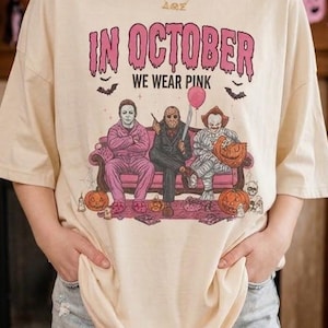 May include: A cream-colored t-shirt featuring the text "IN OCTOBER WE WEAR PINK" in a dripping font. The graphic depicts three horror movie characters sitting on a pink sofa, surrounded by pumpkins, bats, and a pink balloon.