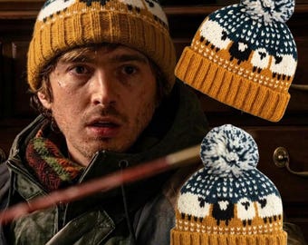 May include: A knit beanie featuring a mustard yellow ribbed cuff, a dark blue and white patterned section with a repeating sheep motif, and a white and blue speckled pom-pom on top. The hat is displayed in two views to show the design.