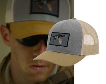 May include: A trucker cap featuring a grey front panel, a tan brim, and tan mesh back panels. A rectangular patch is centred on the front, displaying a detailed graphic of a duck in flight against a dark background, showcasing natural plumage colours.