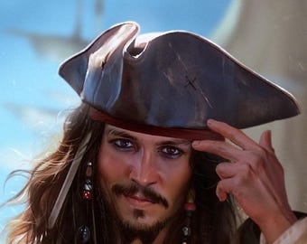May include: A dark brown tricorn hat with a worn texture and a red fabric band. The hat features a small cross mark on the side. The wearer has long brown hair with braided sections adorned with various beads and charms, and dark eye makeup.