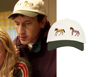 May include: A baseball cap with an off-white crown and a dark olive green brim. The front of the crown features two embroidered horse figures, one wearing a tan blanket and the other wearing a patterned maroon and white blanket, positioned side by side.