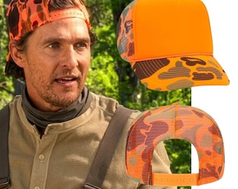 May include: A trucker hat featuring a solid bright orange front panel, a mesh back, and a camouflage pattern in shades of orange and brown on the brim and rear panels. The hat includes an adjustable plastic snapback closure at the rear.