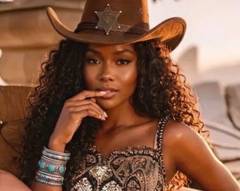 May include: A brown felt cowboy hat featuring a silver-toned six-pointed star badge on the front crown. The hat has a dark brown braided hatband and a wide, curved brim. The accessory is worn with a stack of silver and turquoise bracelets.