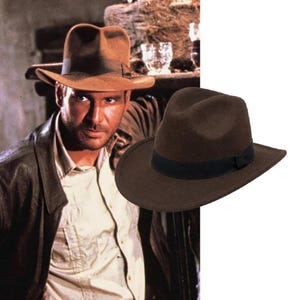 May include: A dark brown felt fedora hat featuring a structured crown with a centre crease and a wide brim. The hat is accented with a solid black grosgrain ribbon band encircling the base of the crown, providing a distinct contrast to the dark brown felt.
