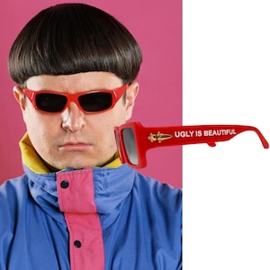 May include: Bright red sunglasses featuring dark lenses and the text UGLY IS BEAUTIFUL printed in white on the temple. The frame includes a gold-colored metallic emblem near the hinge, showcasing a distinct rectangular shape and bold design.