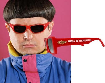 May include: Bright red sunglasses featuring dark lenses and the text UGLY IS BEAUTIFUL printed in white on the temple. The frame includes a gold-coloured metallic emblem near the hinge, showcasing a distinct rectangular shape and bold design.