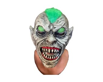 May include: A full-head creature mask featuring a textured grey skin tone, a bright green mohawk, and glowing green eyes. The mask displays a wide, menacing grin with sharp, pointed white teeth set against deep red gums and dark shading.