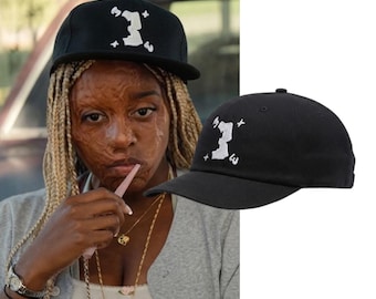 May include: A black baseball cap featuring a white embroidered graphic on the front panel. The design consists of a stylised, blocky letter shape surrounded by four small, four-pointed star symbols. The cap has a structured crown and a curved brim.