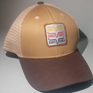 May include: A tan and brown baseball cap with a mesh back and a brown brim. The front features a square patch with the text Kimes Ranch in three stacked lines, each with a different colored line graphic above the brand name.