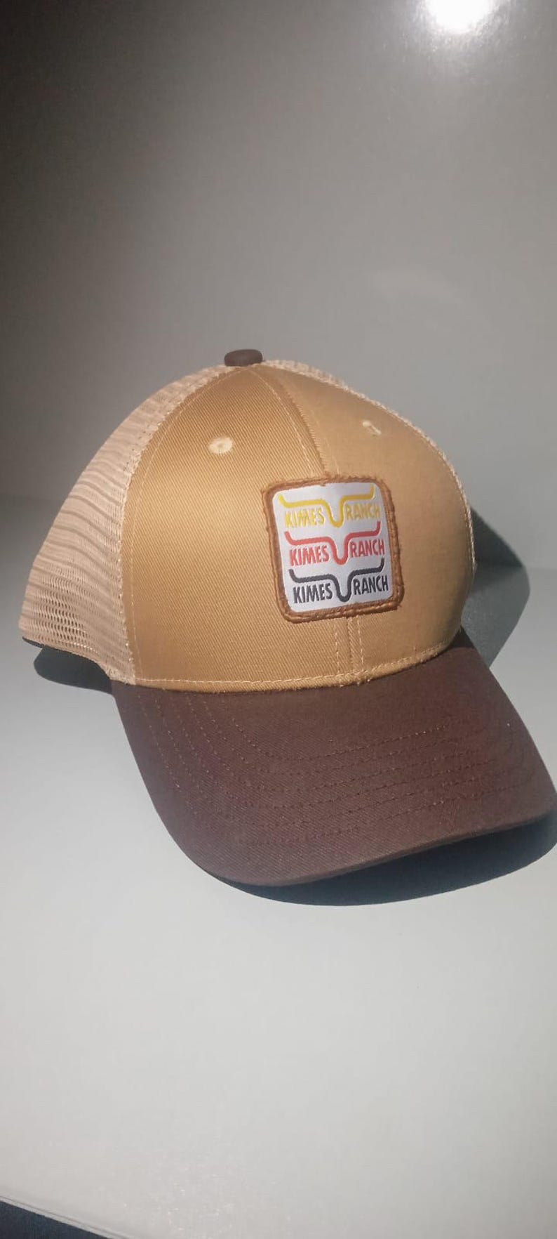 May include: A tan and brown baseball cap with a mesh back and a brown brim. The front features a square patch with the text Kimes Ranch in three stacked lines, each with a different colored line graphic above the brand name.