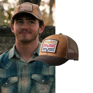 May include: A tan and brown trucker hat featuring a rectangular patch on the front. The patch displays the text "KIMES RANCH" in three stacked lines with a stylized longhorn logo above each line in yellow, red, and black colors respectively.