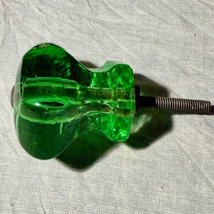 May include: A translucent emerald green glass cabinet knob with a faceted, multi-sided shape. The knob features a central metal bolt with a threaded screw extending from the back, designed for attachment to furniture or cabinetry doors.
