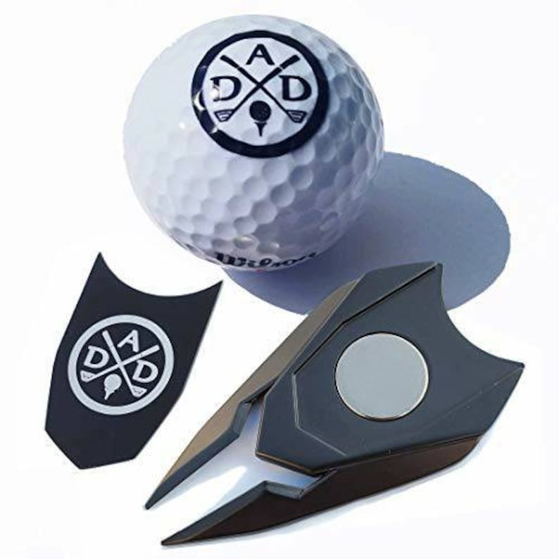 May include: A black golf ball retriever with a magnetic base. The retriever is shaped like a sharp point and has a silver magnetic disc on the top. The retriever is next to a white golf ball with a blue logo that says "A D X D" with crossed golf clubs.