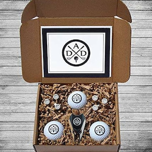 May include: A brown cardboard box with a white lid. The lid has a black and white logo with the letters "A D D" and crossed golf clubs. The box contains a set of golf balls with the same logo, a golf ball retriever, and golf tees.