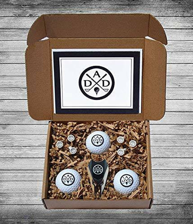 May include: A brown cardboard box with a white lid. The lid has a black and white logo with the letters "A D D" and crossed golf clubs. The box contains a set of golf balls with the same logo, a golf ball retriever, and golf tees.