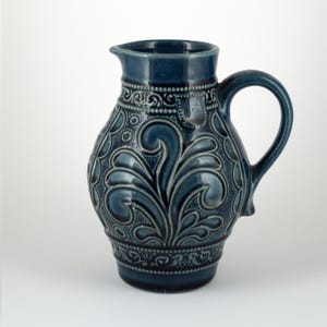 May include: A dark blue ceramic pitcher featuring a glossy finish and intricate embossed scrollwork patterns. The vessel has a rounded body, a curved handle, and a distinct spout, with a small shield-shaped emblem near the top rim.