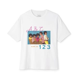 May include: A white short-sleeve t-shirt featuring a graphic print of five individuals sitting on a wooden bench against a blue background. Above the image is the text ABC in pink, and below the image is the text EASY AS 123 in orange and teal.