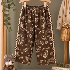 May include: Brown trousers featuring a white paisley and floral pattern with a cream-coloured rickrack trim along the side seams. The trousers have an elasticated waistband and are displayed on a wooden hanger.