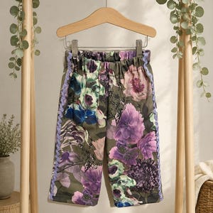 Puede incluir: Pantalones de color verde oliva con un estampado floral vibrante en tonos morado, rosa y azul. Los pantalones tienen una cintura elástica y están decorados con un ribete festoneado de color morado claro en el lateral de cada pierna.
