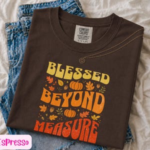 May include: A dark brown short-sleeve t-shirt featuring the text BLESSED BEYOND MEASURE in yellow, orange, and red. The graphic includes illustrations of pumpkins, acorns, and autumn leaves. A thin gold chain necklace rests on the neckline.