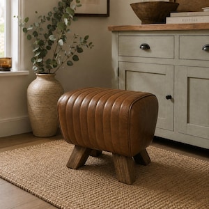 May include: A brown leather ottoman with a ribbed, rounded top surface and four sturdy wooden legs. The piece features a smooth, polished finish and a structured, rectangular shape, providing a stable base for seating or resting feet in a room.