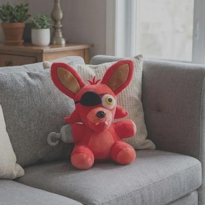 Puede incluir: Un peluche de un personaje con pelaje rojo brillante, orejas grandes y puntiagudas y un parche negro sobre un ojo. La figura tiene un ojo amarillo, una nariz pequeña negra, dos dientes blancos visibles y un gancho gris en el brazo derecho.