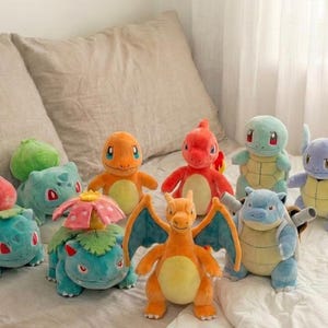 Peut inclure: Un ensemble de neuf peluches représentant diverses créatures, dont un Bulbasaur bleu sarcelle, un Herbizarre vert, un Florizarre à fleur rose, un Salamèche orange, un Reptincel rouge, un Dracaufeu ailé, un Carapuce bleu clair, un Carabaffe et un Tortank.