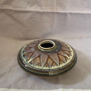 May include: A low-profile, circular ceramic vase featuring a central opening and a radial, leaf-like embossed pattern. The glaze displays a blend of earthy brown, muted green, and cream tones with a glossy finish across the entire exterior surface.