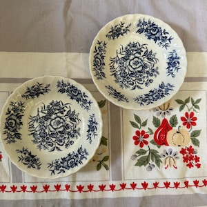 May include: Two round ceramic bowls with scalloped edges, featuring a white base color and a detailed dark blue floral pattern. The design includes a central bouquet of flowers surrounded by smaller floral motifs along the inner rim of each bowl.
