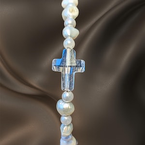 May include: A beaded bracelet featuring a series of irregular white freshwater pearls of varying sizes interspersed with small round white beads and a central clear glass cross charm with a vertical ribbed texture and a polished finish.
