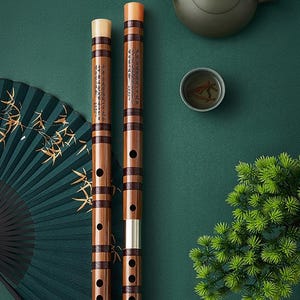 May include: Two cylindrical bamboo flutes with dark brown binding bands and light-coloured end caps. The flutes feature black ink calligraphy characters printed vertically along the wood grain and multiple circular finger holes along the length.