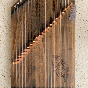 May include: A wooden guzheng instrument featuring a dark brown finish with a natural wood grain pattern. The instrument has multiple white strings stretched across a series of small, stepped wooden bridges and includes a decorative fan engraving.