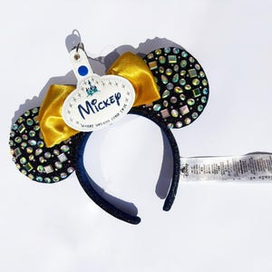 May include: A headband featuring two black ears covered in iridescent rhinestones, a gold satin bow, and a white oval tag with the text "Mickey" and "Where dreams come true" printed in blue, along with a small blue dot and a Disney logo tag.
