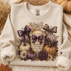May include: A cream-colored crewneck sweatshirt featuring a graphic of a coffee cup with latte art, surrounded by pumpkins, purple flowers, and striped bows. The text on the cup reads Autumn COFFEE & Cozy Mood in dark brown lettering.