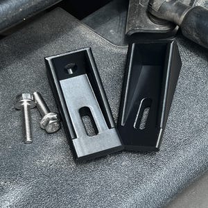 May include: Two black, wedge-shaped metal brackets with a rectangular slot and a circular hole, accompanied by two silver-colored metal bolts with hexagonal heads and threaded shafts, designed for mechanical mounting or adjustment purposes.