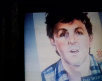 May include: A portrait print of Paul McCartney featuring dark hair and a blue plaid shirt. The image includes a circular red stamp with the number 32 and the text Paul McCartney printed in a dark rectangular box at the bottom right corner.