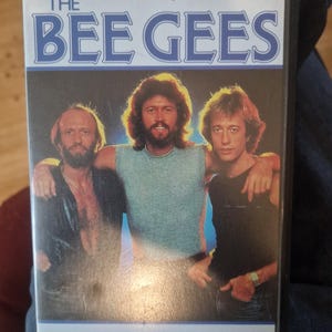 Könnte beinhalten: Eine VHS-Kassette mit dem Titel The Story of The Bee Gees, die ein Foto von drei Männern zeigt, die ihre Arme umeinander gelegt haben. Das Cover enthält den Text The Story of The Bee Gees sowie die Logos von CCTV, Videophonic Sound und MGM/UA.