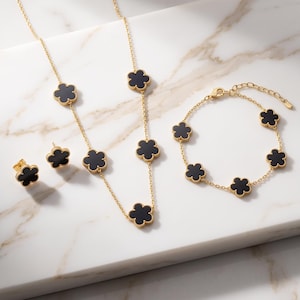 May include: A gold-toned jewelry set consisting of a long necklace, a bracelet, and a pair of stud earrings. Each piece features multiple black clover-shaped charms with gold-toned edges, connected by a delicate gold-toned chain link design.