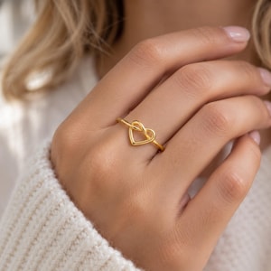 May include: A gold-toned ring featuring a delicate, continuous wire design that forms a stylized heart shape. The band is thin and polished, creating a smooth, minimalist silhouette that sits centered on the finger with a bright, reflective finish.