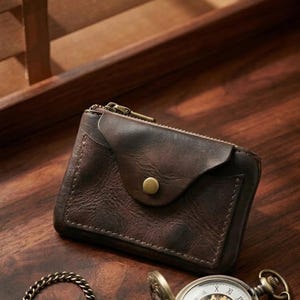 Peut inclure: Un portefeuille en cuir marron foncé doté d'un compartiment supérieur zippé et d'une poche frontale à rabat fermée par un bouton-pression couleur laiton. À côté se trouve une montre de poche argentée avec mécanisme apparent et chaîne en métal.