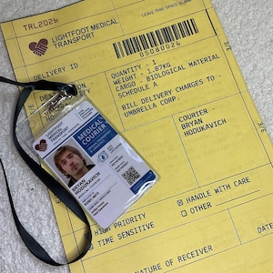May include: A yellow Lightfoot Medical Transport delivery form with a barcode and text including Cargo - Biological Material, placed under a clear plastic ID badge for Medical Courier Bryan Hodukavich with a black lanyard and a QR code.