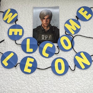 Op de afbeelding: Een decoratieve slinger bestaande uit blauwe ronde kartonnen schijven met gele letters die WELCOME LEON vormen, geregen aan een zwart koord. Boven de tekst bevindt zich een rechthoekig portret van een man in een tactisch vest met R.P.D.