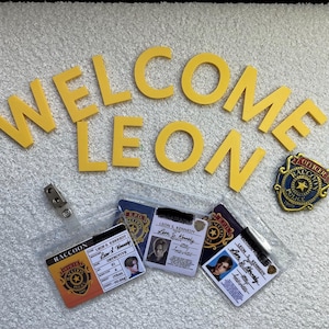 以下が含まれることがあります： 「WELCOME LEON」と綴られた黄色のプラスチック文字が、ラクーン市警の身分証3枚と金色の金属製警察バッジの上に配置されています。身分証にはレオン・S・ケネディの名前、写真、詳細情報が印刷されています。