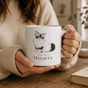 May include: A white ceramic mug featuring a black and white illustration of a fluffy cat sitting down. Below the cat, the text reads "TINY BUT MIGHTY" in a clean, serif font. The mug has a standard handle and a smooth, glossy finish.