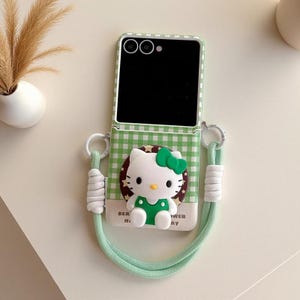 Puede incluir: Una funda para teléfono plegable con estampado de cuadros vichy en verde y blanco, que presenta una figura tridimensional del personaje Hello Kitty con lazo y traje verde, unida a un cordón de cuerda verde claro con detalles blancos.