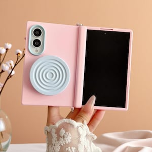 May include: A light pink foldable smartphone case featuring a light blue camera module surround and a circular light blue magnetic grip attachment with a concentric ring pattern on the back panel, paired with a matching light pink frame.