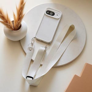 May include: A white smartphone case with a matching white fabric crossbody strap attached by silver-toned metal lobster clasps. The case features a raised camera cutout area and a smooth, solid white back surface, providing a clean and functional design.